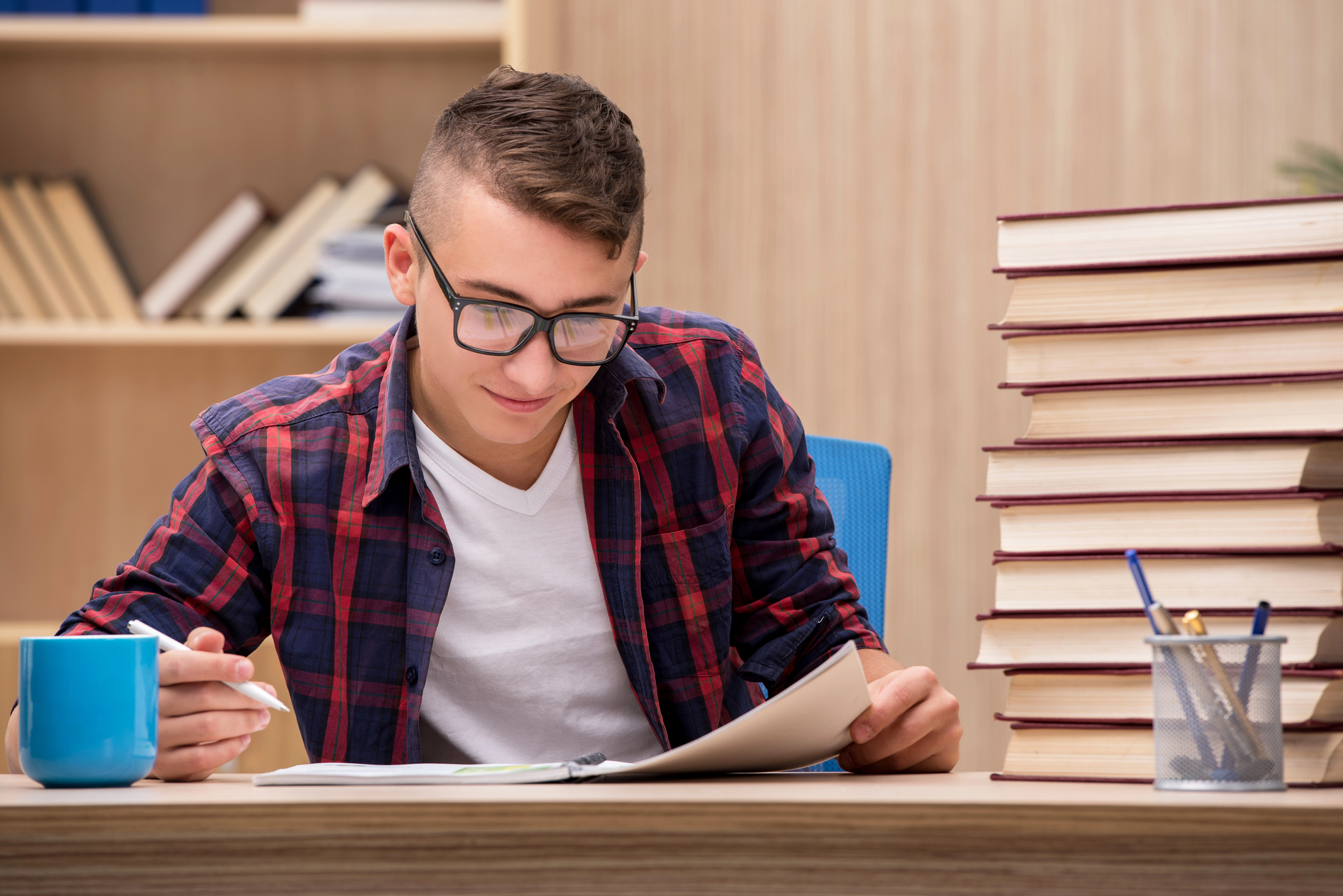 High school boy writing at home desk with stack of books nearby.