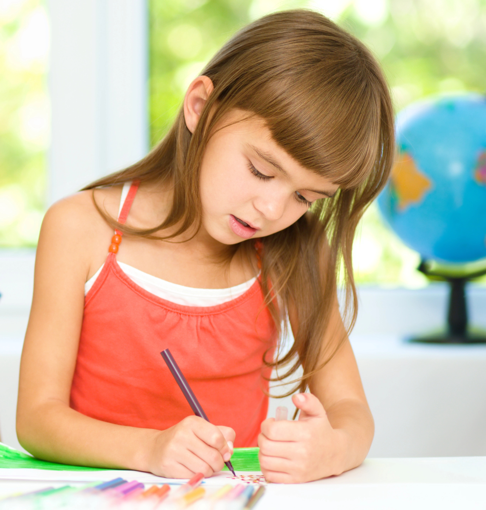 Young girl with brown hair intently writing in front of window.